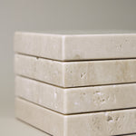 Stack of square MAIE coasters in Travertino Classico VC, showing beige veining, natural pores and tonal variation.