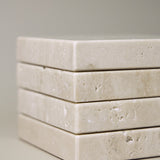 Stack of square MAIE coasters in Travertino Classico VC, showing beige veining, natural pores and tonal variation.