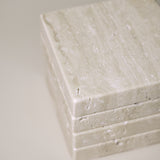 Stack of square MAIE coasters in Travertino Classico VC, showing beige veining, natural pores and tonal variation.