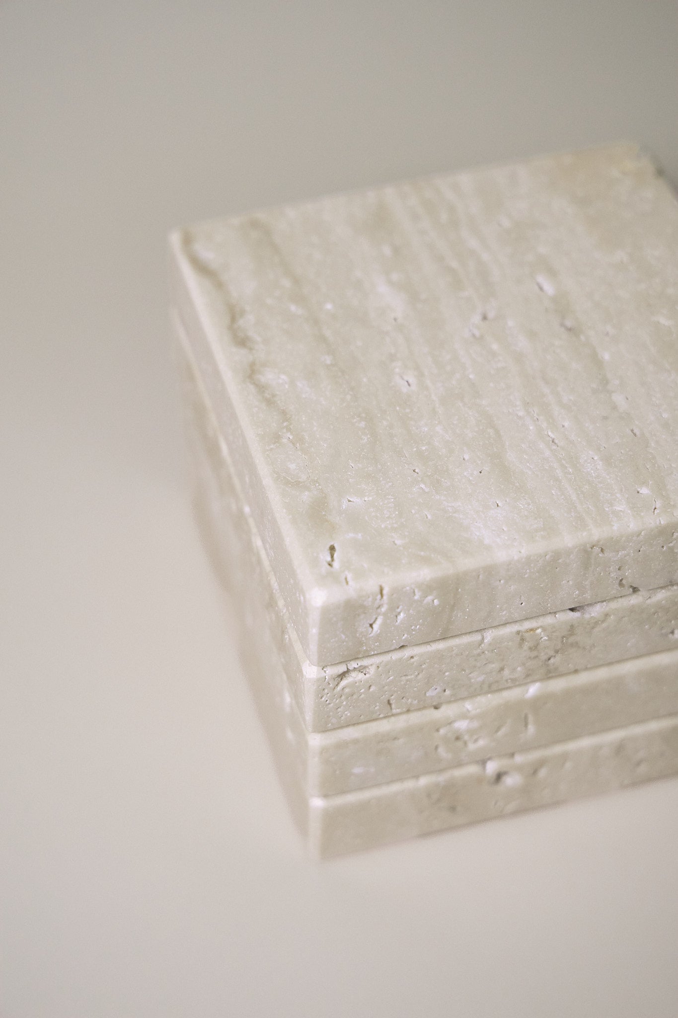 Stack of square MAIE coasters in Travertino Classico VC, showing beige veining, natural pores and tonal variation.
