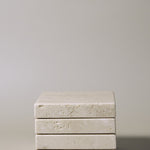 Stack of square MAIE coasters in Travertino Classico VC, showing beige veining, natural pores and tonal variation.