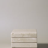 Stack of square MAIE coasters in Travertino Classico VC, showing beige veining, natural pores and tonal variation.