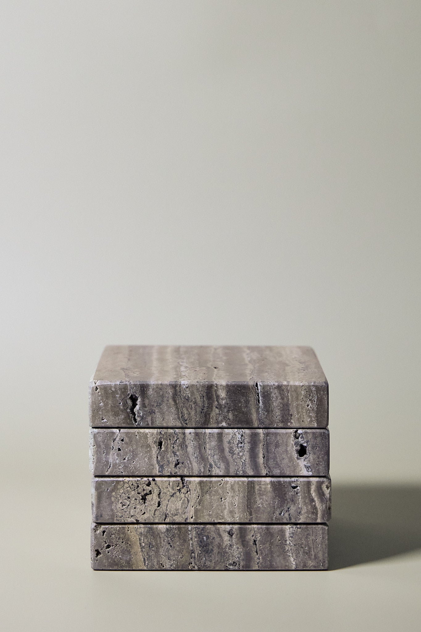 Stack of square MAIE coasters in Travertino Marrone, showing layered grey-brown veining, natural pores and straight edges.
