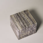 Stack of square MAIE coasters in Travertino Marrone, showing layered grey-brown veining, natural pores and straight edges.