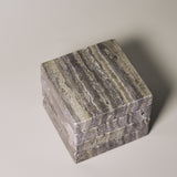 Stack of square MAIE coasters in Travertino Marrone, showing layered grey-brown veining, natural pores and straight edges.